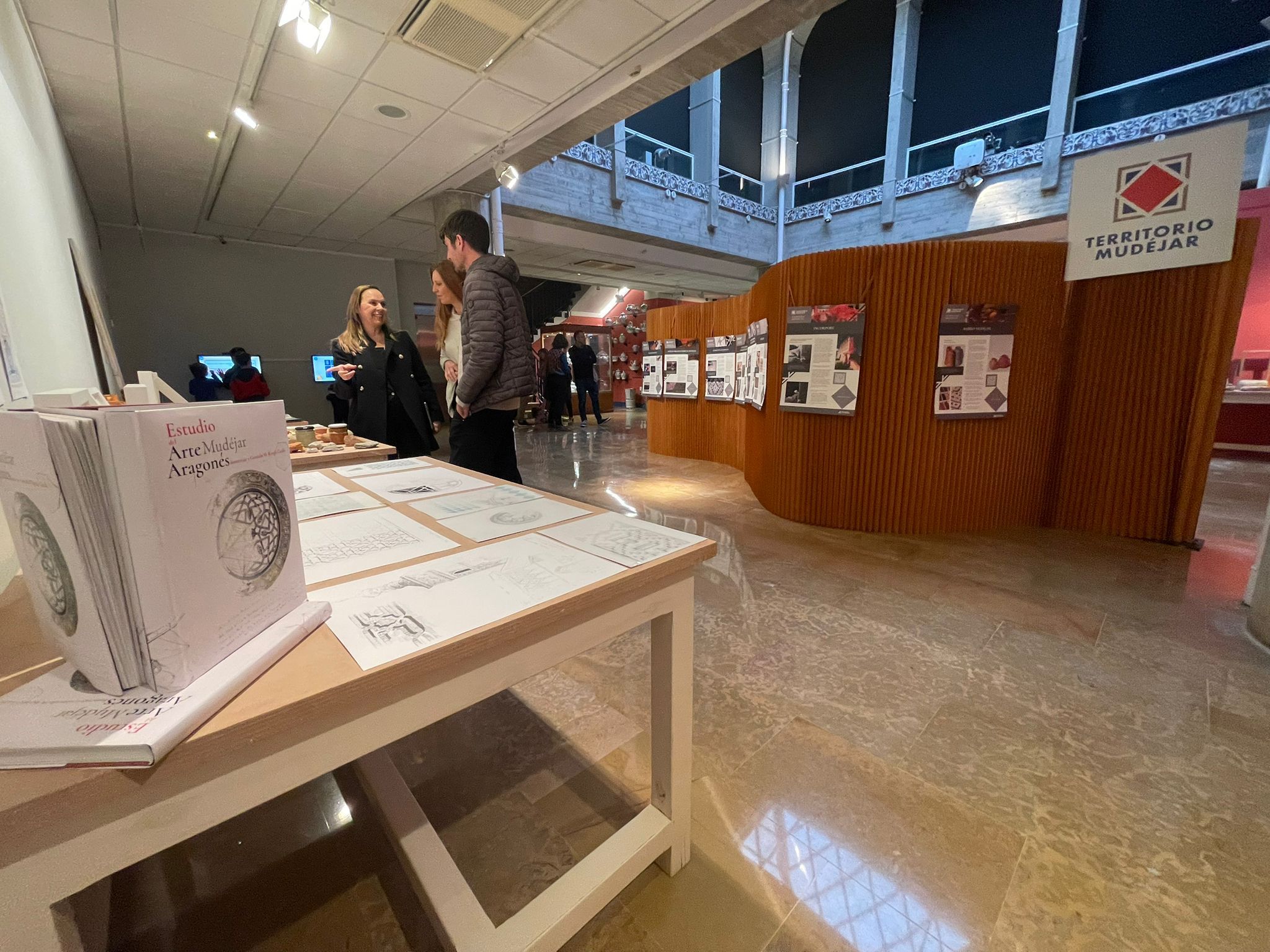 Vista de la exposición "Estrategias y proyectos de Territorio Mudéjar" en Muel, con visitantes conversando junto a paneles y publicaciones.