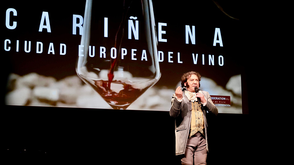 Presentador en el escenario durante el acto oficial de proclamación de Cariñena como Ciudad Europea del Vino 2025, con una gran copa de vino proyectada al fondo.