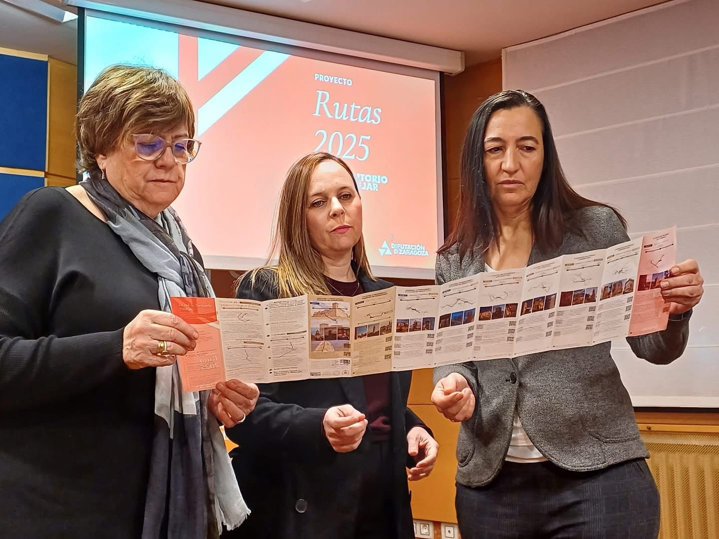Tres mujeres presentan el nuevo folleto de las Rutas Territorio Mudéjar 2025 en un acto institucional con proyección de fondo.